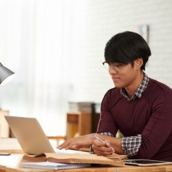 Vietnamese college student reading information on laptop and taking notes